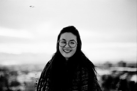 A black and white headshot of the author, an Asian American woman with long brown hair and geometric shaped glasses, with a shawl from Dharamshala wrapped around her.