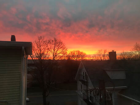 This is a picture of a sunrise from a third floor bedroom window in Northampton, Massachussetts. The sunrise is neon orange, pink, and yellow. Telephone lines and roofs are on the horizon. 
